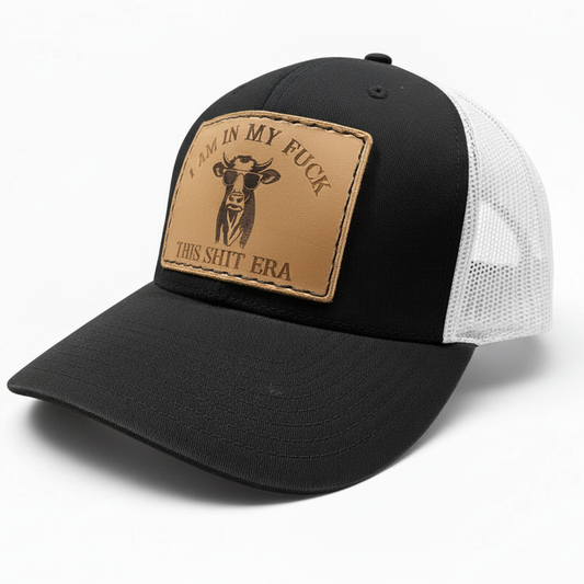 Black trucker cap with a brown leather patch featuring text and a cow illustration on a white background.