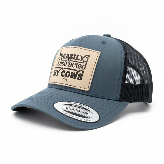 Grey and black cap with a patch reading 'Easily Distracted by Cows' on a whitesurface