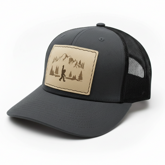 Gray trucker cap with a tan leather patch featuring a mountain and hiking design on a white background