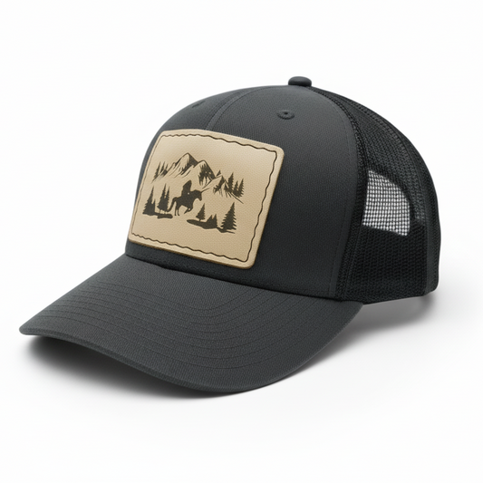 Black trucker cap with a beige patch featuring mountains and trees on a light gray background