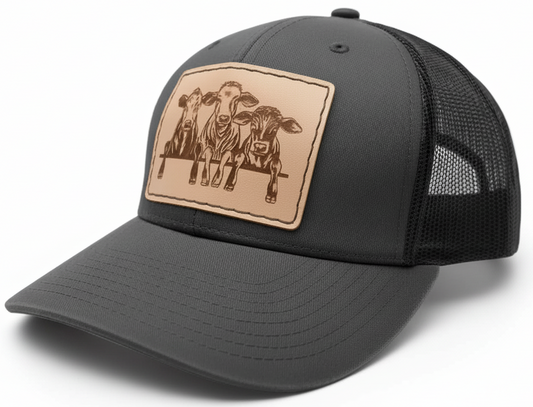 Black and grey trucker cap with a tan leather patch featuring cow designs on a white background