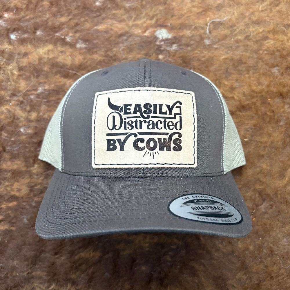 Gray cap with a patch reading 'Easily Distracted by Cows' on a brown background
