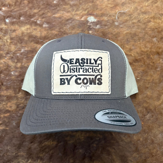 Gray cap with a patch reading 'Easily Distracted by Cows' on a brown background