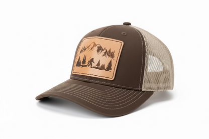 A brown and beige fishing cap with a Sasquatch patch on the front featuring mountain and tree graphics.