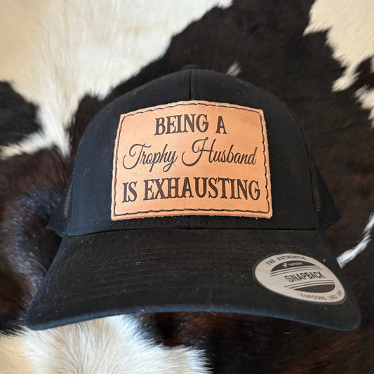 Black cap with a leather patch reading 'Being a Trophy Husband is Exhausting' on a cowhide rug background.