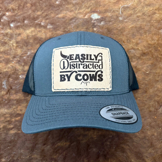 Blue cap with a patch reading 'Easily Distracted by Cows' on a brown surface