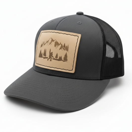 Gray trucker cap with a leather patch featuring a mountain and forest design on a white background