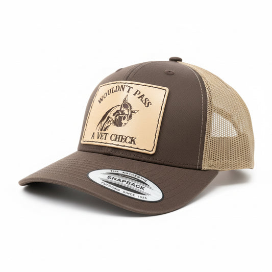Brown cap with a patch reading 'Wouldn't Pass A Vet Check' on a wooden surface.
